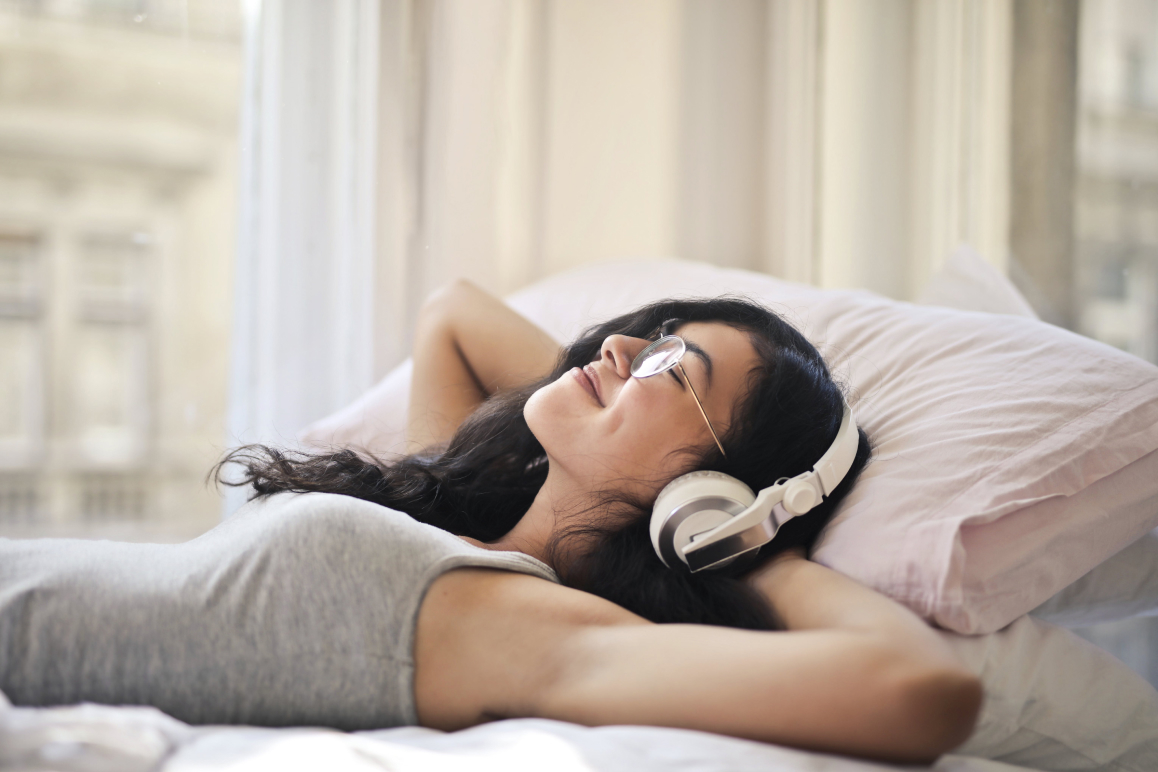 Eine Frau mit dunklen Locken und Brille liegt mit unter dem Kopf verschränkten Armen auf einem Kissen. Ihre Augen sind geschlossen, sie hört Musik mit großen Kopfhörern und lächelt glücklich.