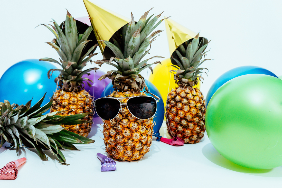 Drei Ananas mit Partyhüte und Sonnenbrillen. Im Hintergrund befinden sich bunte Luftballons.