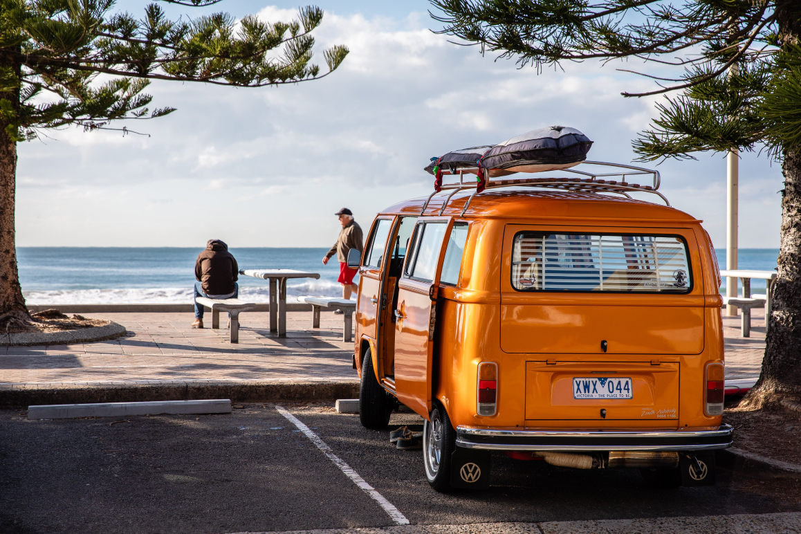 Ein oranger VW Bus steht auf einem Parkplatz vor dem Meer.