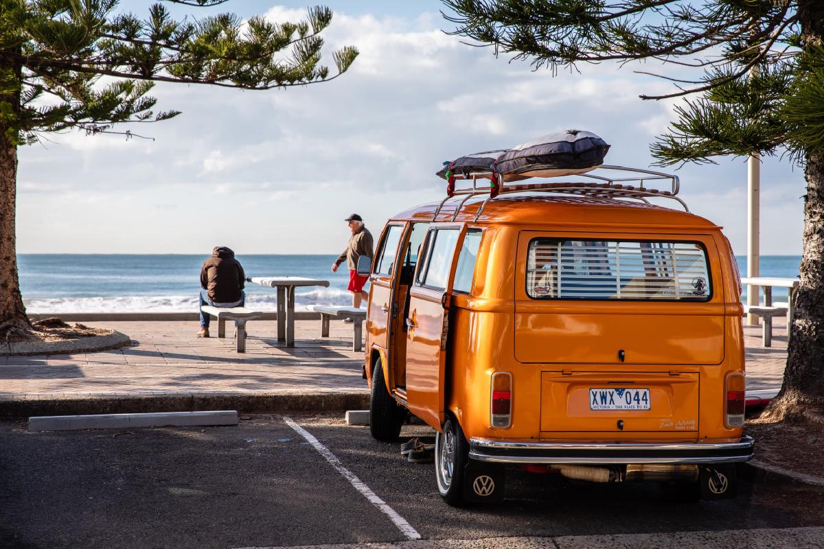 Ein oranger VW Bus steht auf einem Parkplatz vor dem Meer.