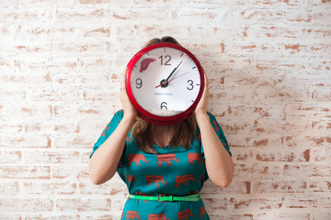 Eine Frau, die sich eine Wanduhr vor ihr Gesicht hält.