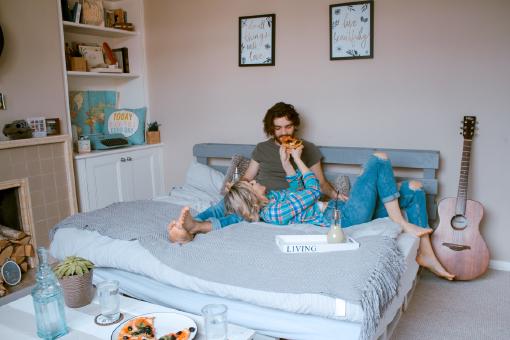 Ein Mann und eine Frau liegen am Bett in ihrer ersten eigenen Wohnung und essen Pizza. Die Wohnung ist über eine Haushaltsversicherung versichert.