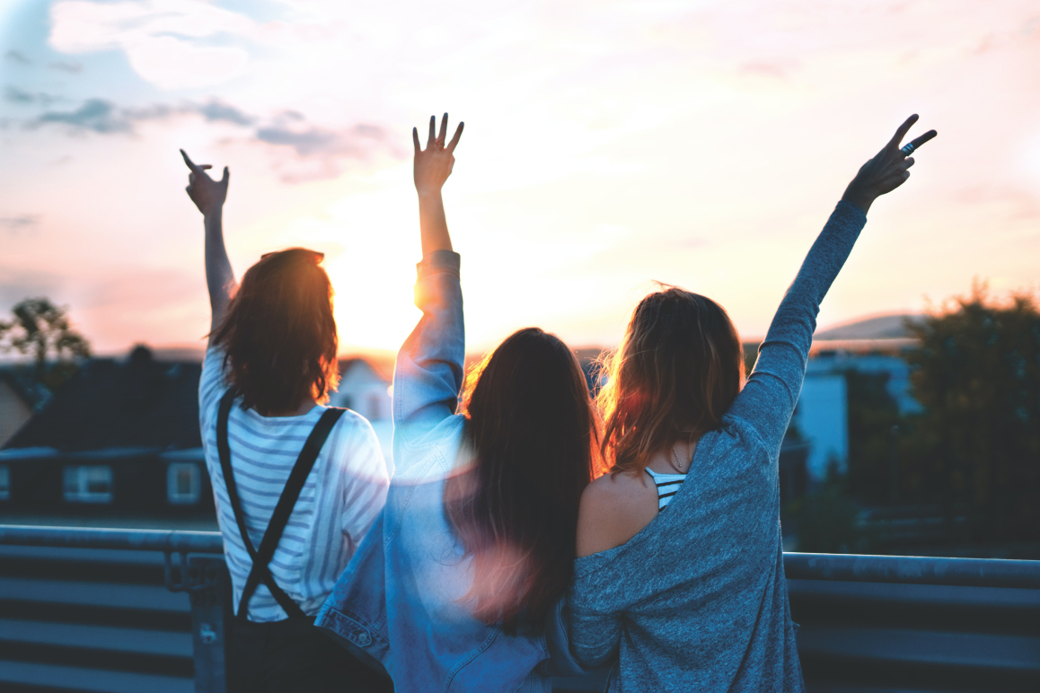 Drei Frauen, die ihre Hände in die Luft halten. Im Hintergrund geht die Sonne unter.