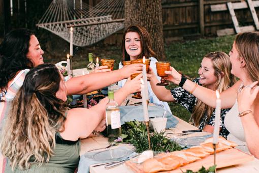 Fünf lächelnde Frauen sitzen an einem Tisch und stoßen mit ihren Gläsern an.